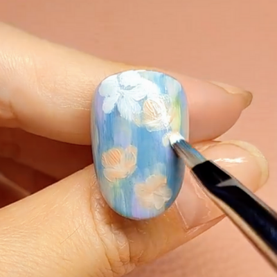 Close-up of a hand holding a nail with blue base color and floral designs being applied with a brush.