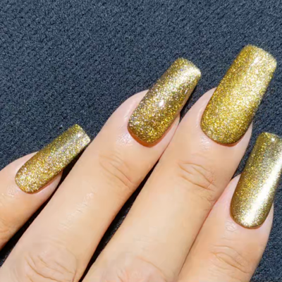 Hand with long nails painted in glittery gold nail polish on a black textured surface.