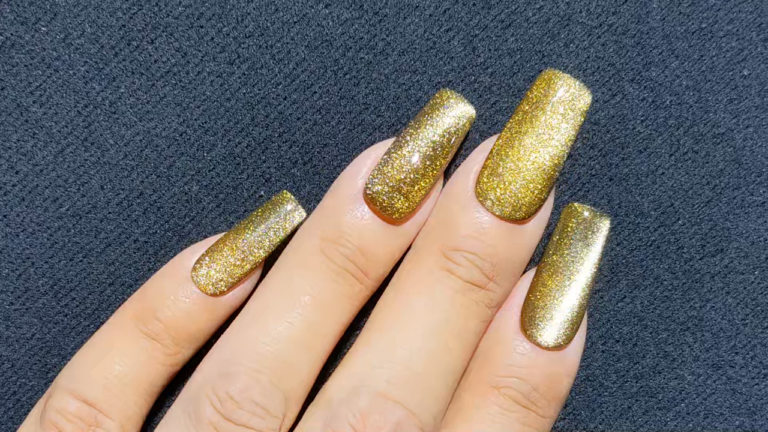 Hand with long nails painted in glittery gold nail polish on a black textured surface.