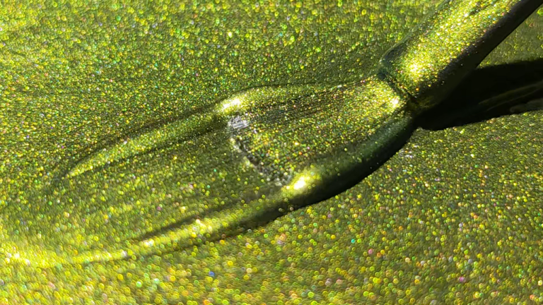 Close-up view of a brush applying glittery green cat eye gel polish with holographic effects.